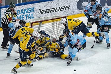 Intense ice hockey match with players in yellow and blue jerseys battling for the puck near the goal, surrounded by action.