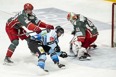 Hockey action as a player attempts a goal against the goalie, with defenders in pursuit on an ice rink.
