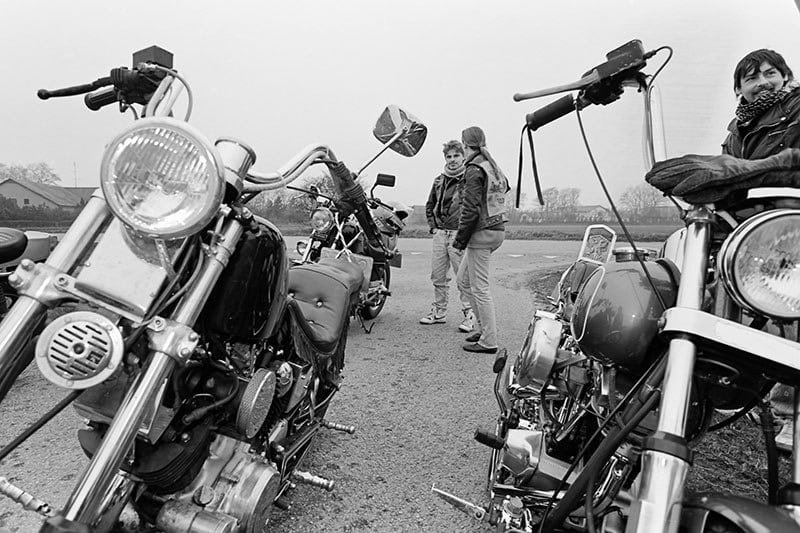 Group of bikers gather near parked motorcycles on a rural road, enjoying a casual meeting on a cloudy day.