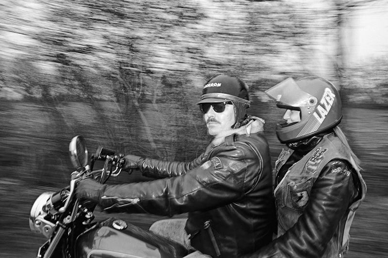 Two motorcyclists ride through a blurred background. Both wear helmets and leather jackets for protection and style.