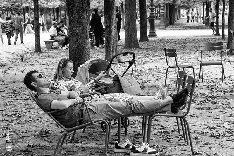 Couple relaxing in park chairs under trees, stroller nearby. Man rests with feet up, woman checking her phone.