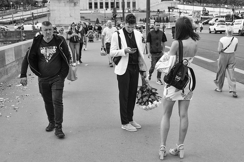 Black and white city street scene with people walking. A man checks his phone, holding flowers; a woman stands nearby.