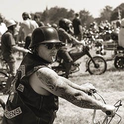 Biker wearing shades and helmet smokes while sitting on motorcycle at an outdoor event with other riders gathered around.