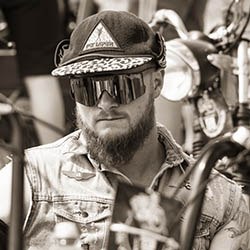 Bearded biker wearing sunglasses and cap sits on motorcycle, capturing a rugged and stylish retro vibe.