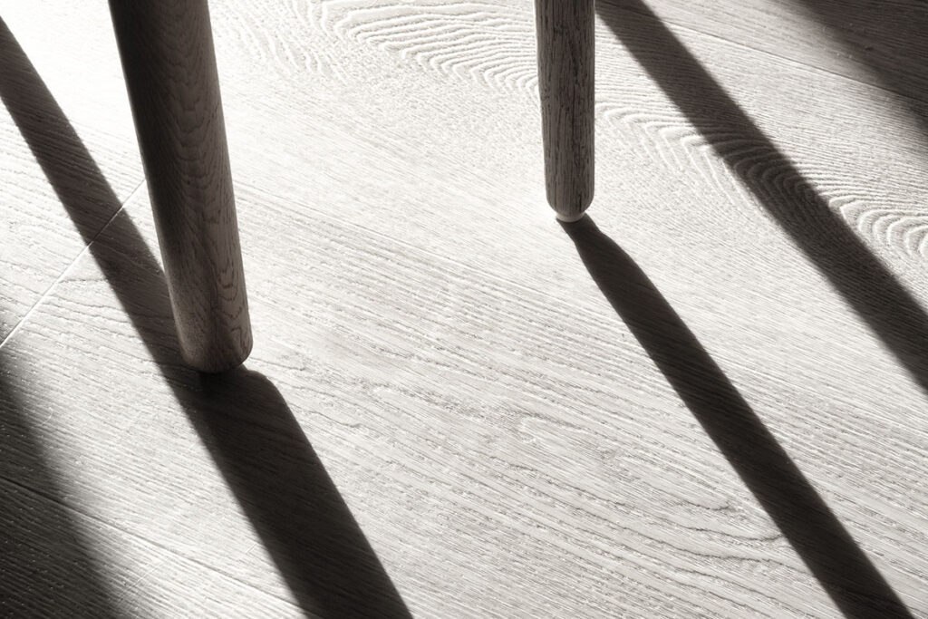 The image showcases shadows of chair legs on a textured wooden floor, highlighting the play of light and shadow patterns.