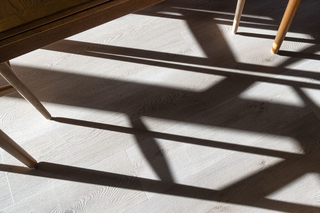 Shadows of wooden chairs cast intriguing patterns on a textured light wood floor, creating an artistic play of light.