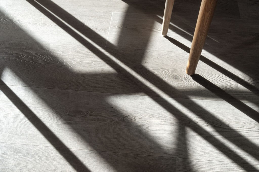 Sunlight casts long, defined shadows of a wooden chair on a textured parquet floor, highlighting intricate details.