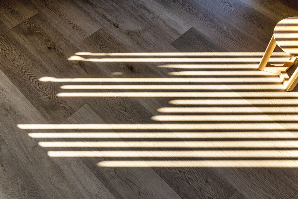 Sunlight streams through blinds, casting striped shadows across a wooden floor with a small stool in the corner.