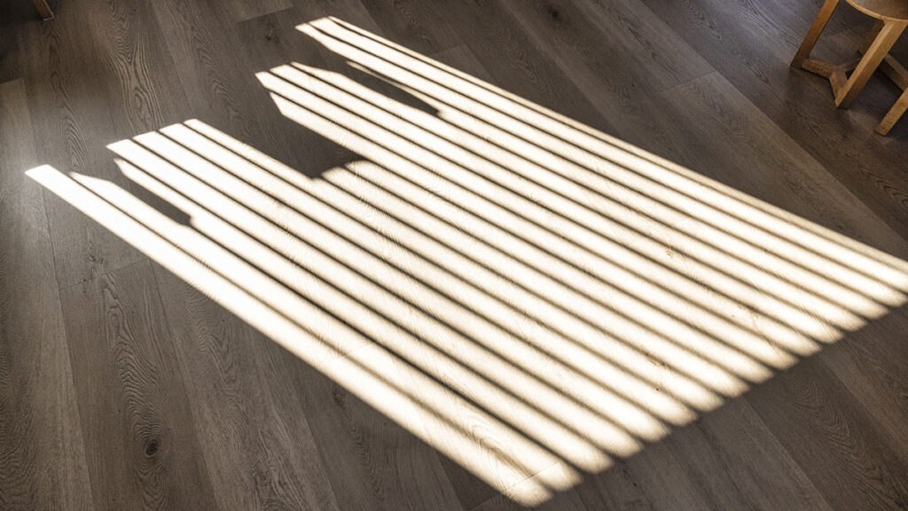 Sunlight filters through blinds, casting long, parallel stripes on a wooden floor, creating a striking pattern.